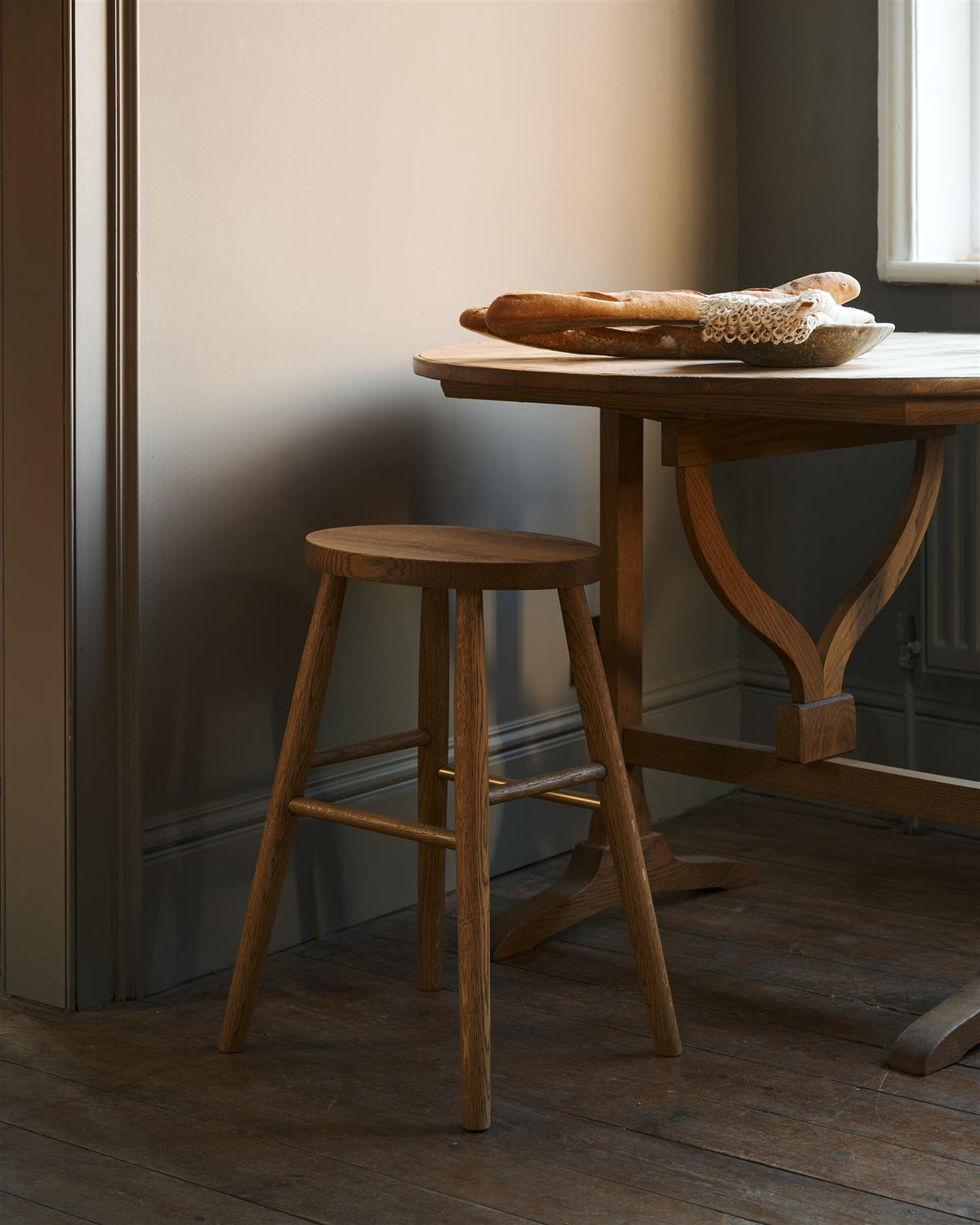 An oblique view of a simple oak milking stool with an oval-shaped seat and a brass footrail by a round pine table in a dark room. TEST