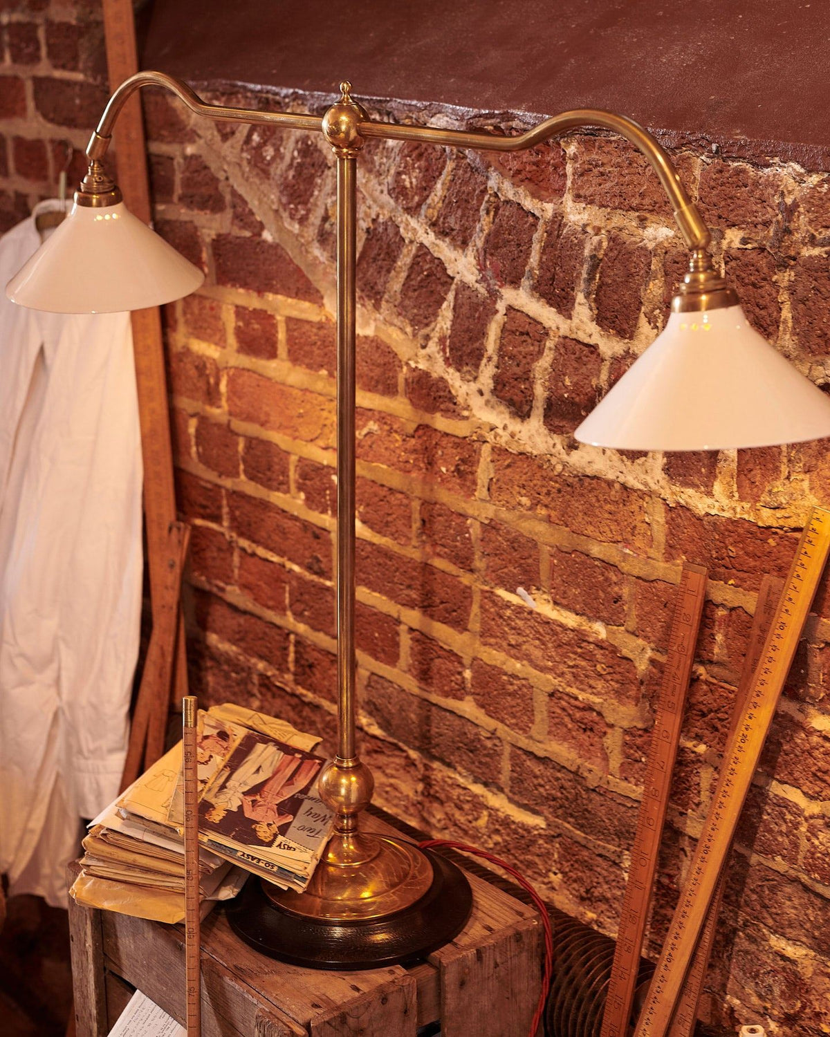 An oblique view of a Victorian-inspired desk lamp with a solid brass foot, stand, two curved arms and two small cream glazed ceramic light shades on a wooden box in front of a red brick wall. TEST
