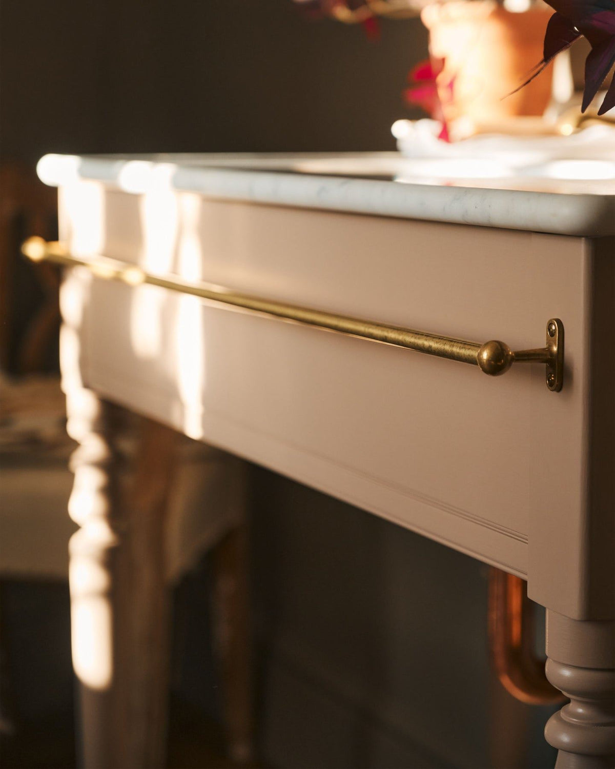 A detailed view of an aged brass towel rail attached to the front of a soft pink painted Victorian-inspired washstand. TEST