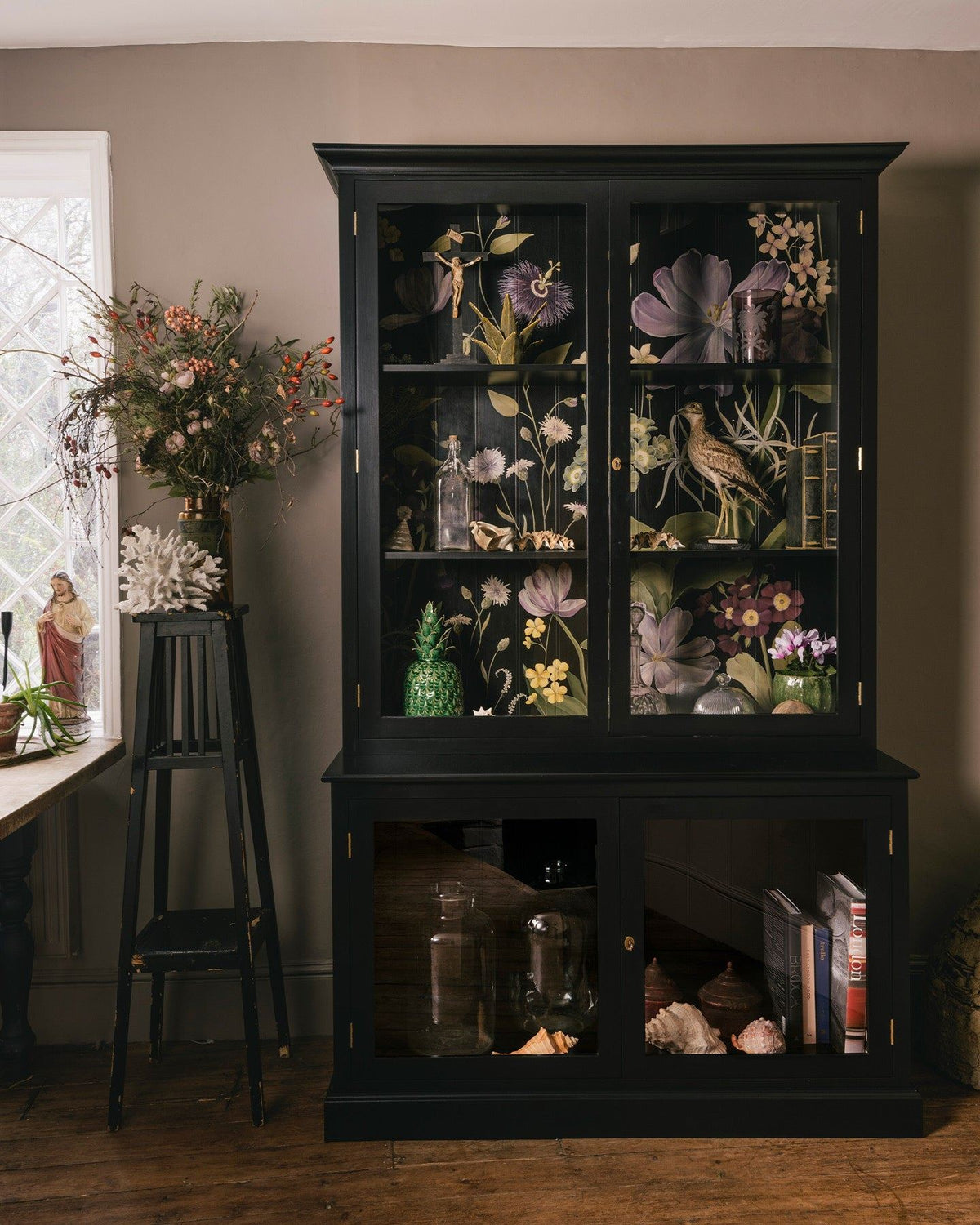 A tall museum-style display cabinet with glazed doors, painted black inside and out and with an elegant and bustling painted floral design on the tongue and groove back panel. TEST