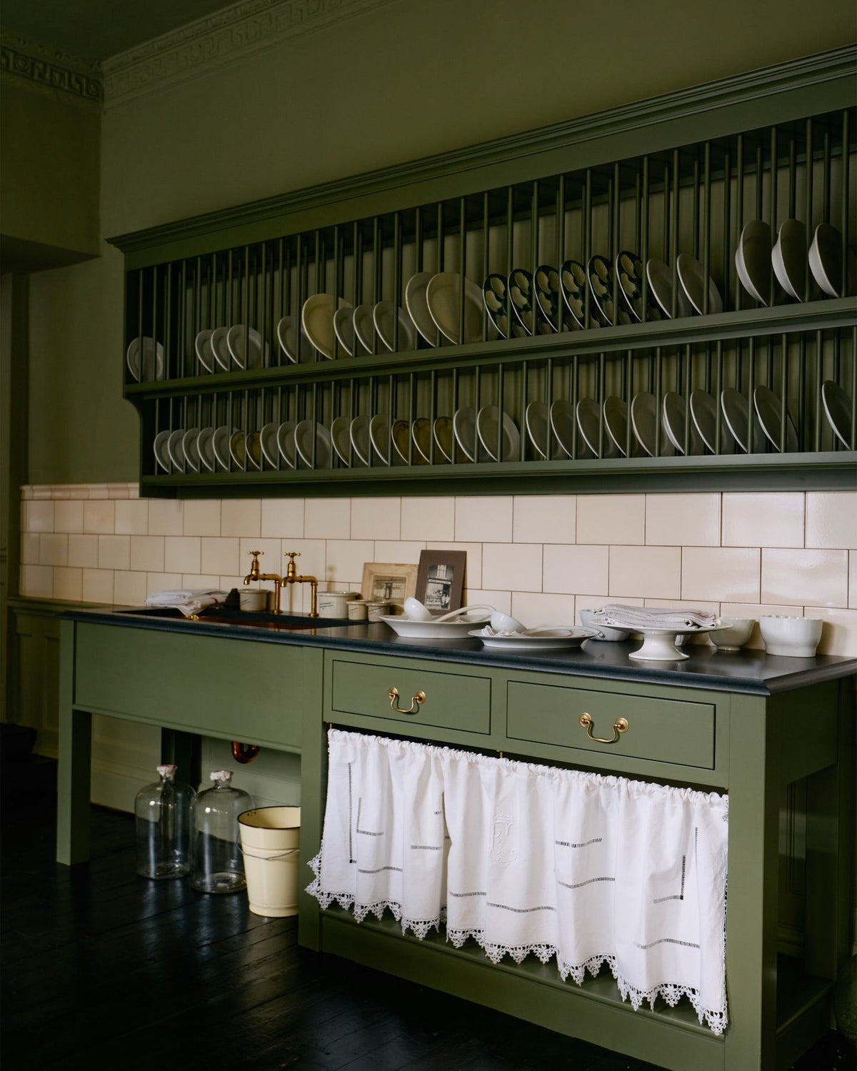 An oblique view of a mossy green painted Victorian-style sink cupboard with a polished brass sink, aged brass bibcock taps and cream subway wall tiles below a traditional wall-mounted plate rack. TEST