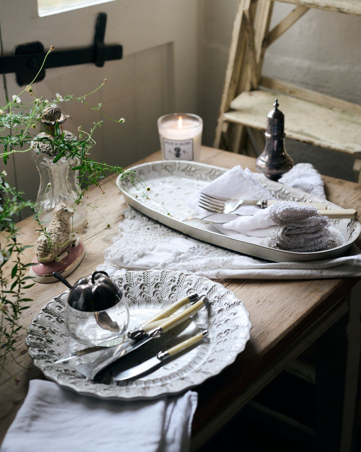 Two bohemian-style white ceramic platters, one oval-shaped, the other round, on a rustic farmhouse table with vintage silverware, white linens and a lit scented candle. TEST