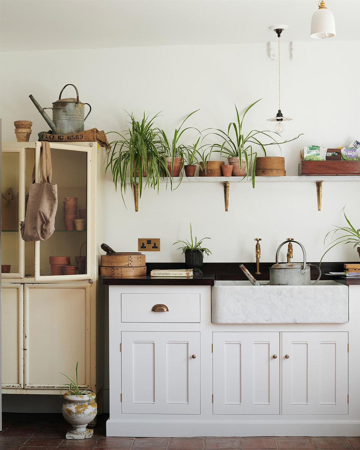 A straight-on view of a classic 13 amp twin plug socket in a mottled aged brass finish in a Mediterannean-inspired kitchen with a white painted Georgian-style sink cabinet, dark hardwood worktop, Carrara marble sink and a shelf full of lush spider plants. TEST