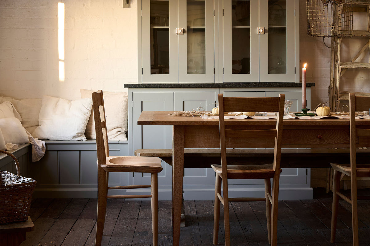 A straight-on view of a rustic kitchen with worn floorboards, white brick walls and grey painted cabinetry. A large table is surrounded by mid-century-style dining chairs with sculpted seat pans.