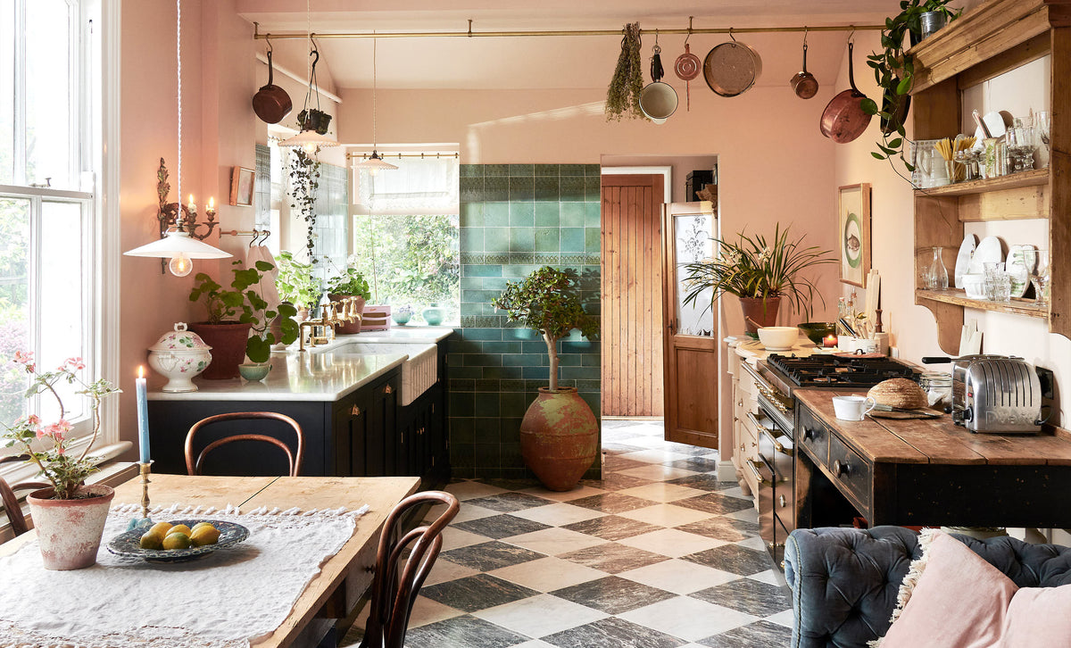 A bright peach-coloured room with a chequerboard marble floor and an eclectic collection of vintage furniture, including a table and chairs, a prep table and a decorative set of drawers. In the left corner is a modest sink run with black painted Classic English cupboards, a Carrara marble worktop and brass fittings. Pots and pans hang from a brass ceiling rail, mixed with a collection of vintage milk glass pendant lights.