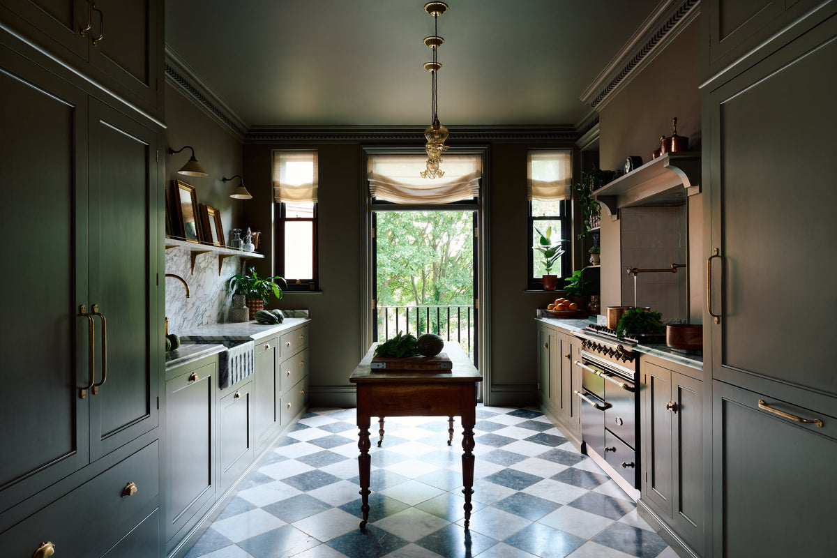 A grand and atmospheric olive green classic English kitchen features a marble worktop and splashback along the sink run, with a single marble sink and an aged brass Ionian tap. A cooker run with an over mantle is paired with an antique prep table on castors. Grand chequerboard marble tiles lead out onto a balcony overlooking green trees.