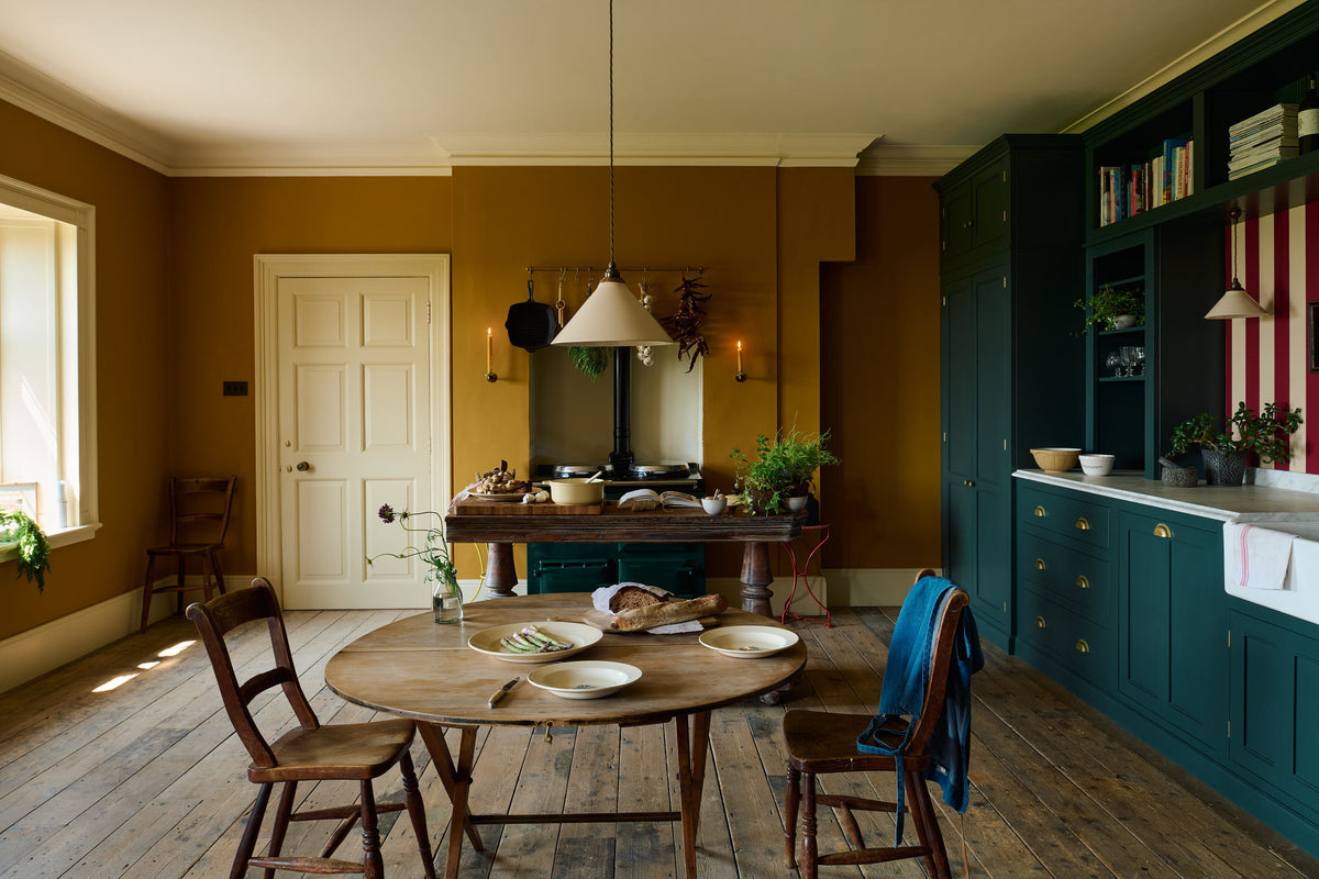 A lovely modern kitchen that retains the charm of its original structure and flooring, using the three primary colours in a muted, mature palette. The spacious kitchen features a Clerkenwell Blue full-height pantry cabinet, a long sink run with a marble worktop, and an open countertop cabinet and shelf. A black gas stove sits within the chimney breast, accompanied by a deVOL cream pendant light above a vintage round wooden table, with matching chairs and a prep table.