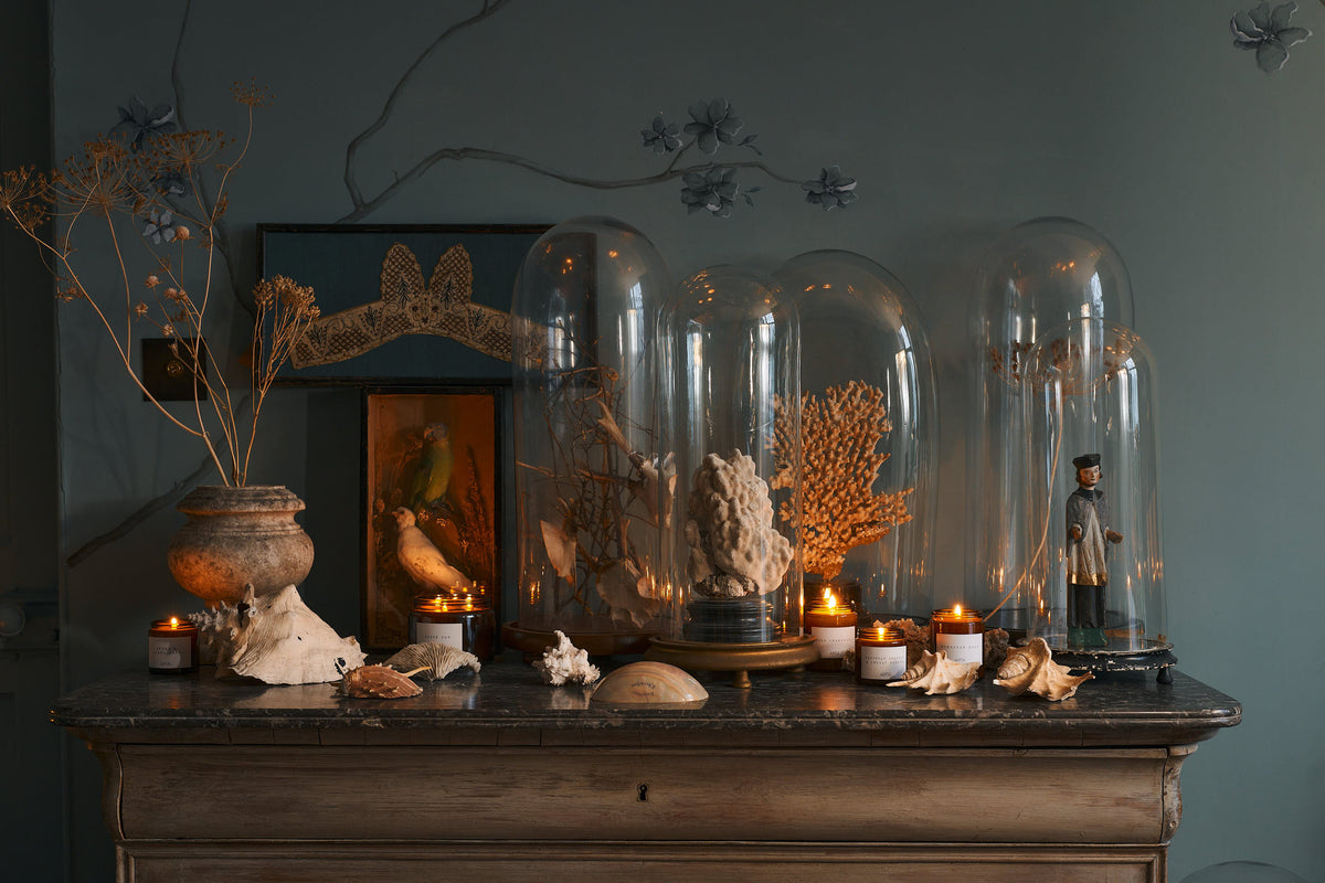 A collection of scented candles in brown glass jars with black lids on a vintage dresser decorated with an assortment of vintage and collectable items.