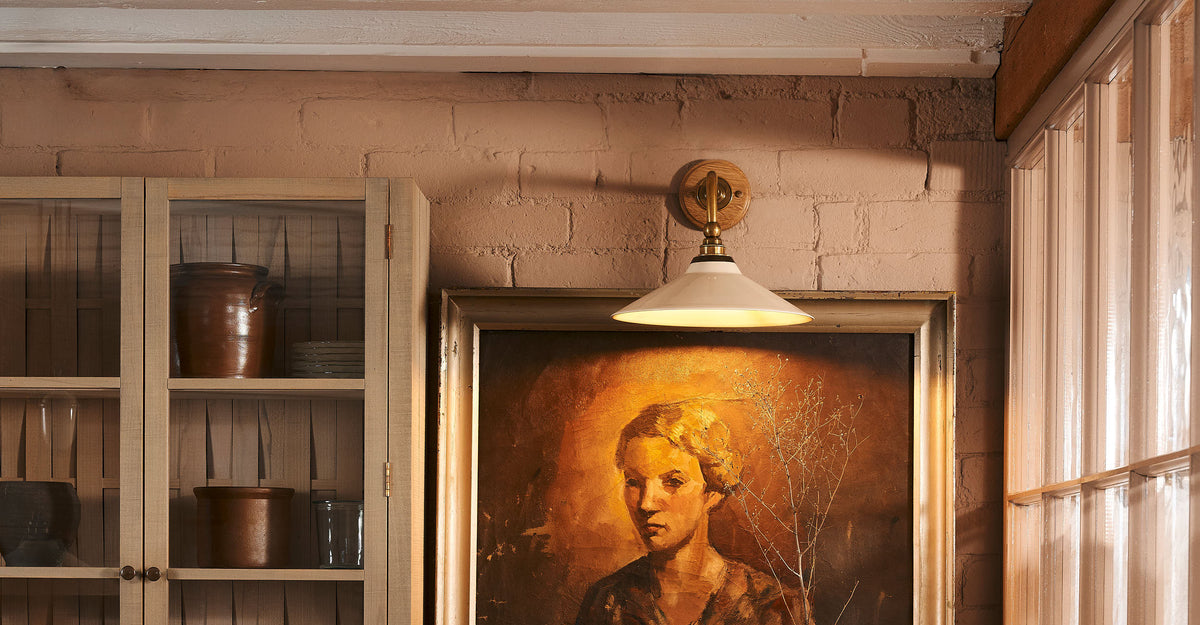 A close-up shot of the Heirloom Task light, which is on the muted pink walls. On the left, a wooden cupboard with a glass door displays bowls and cups. On the right, beneath the wall light, a framed painting of a woman hangs on the wall, its illumination creating an art gallery-like atmosphere.