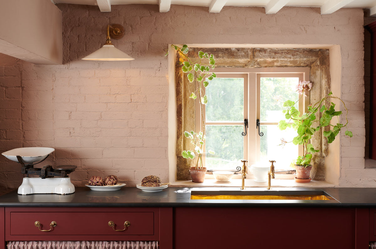A refectory-red Heirloom kitchen’s sink cabinet reveals a polished brass sink, aged brass Mayan taps, café curtain rails, a red and white striped curtain, a plate rack, and an Heirloom dairy table. Beautifully decorated with Heirloom task light, and a vintage scale, this kitchen offers a modern twist on vintage furniture, creating a classic range designed to last.