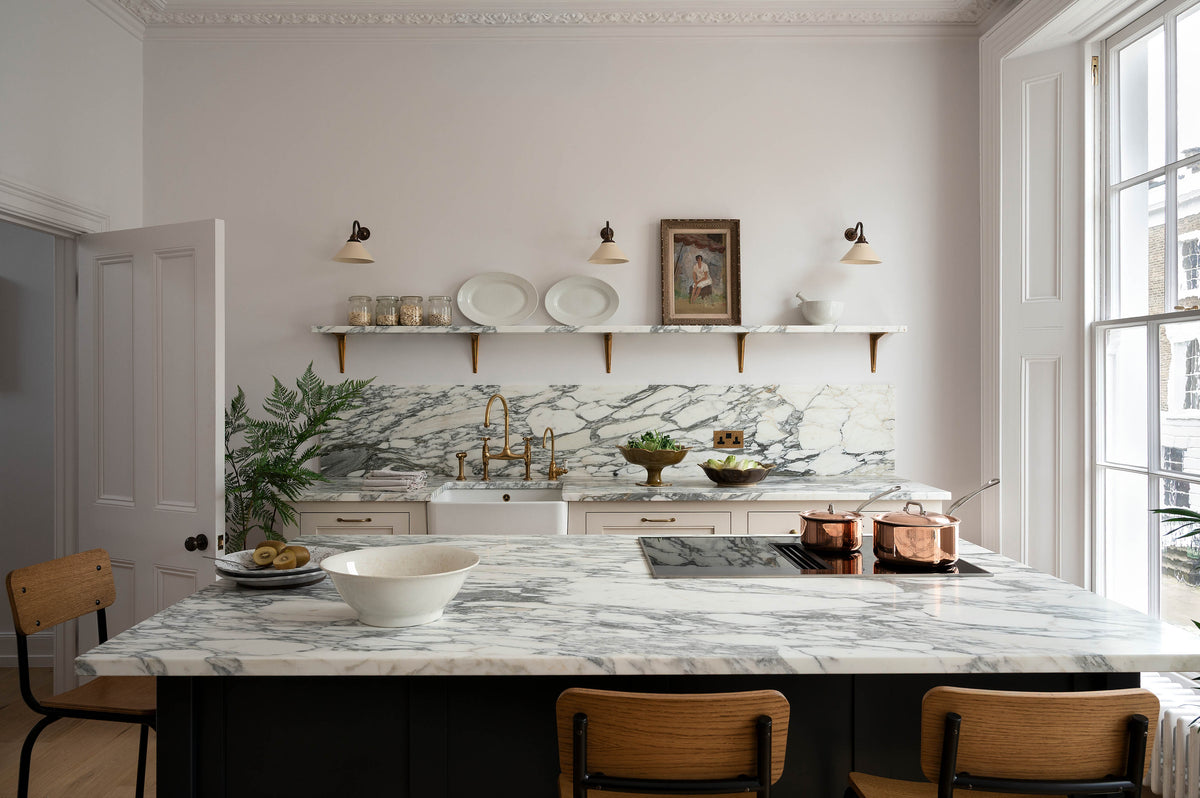 A humble yet elegant off-white Classic English kitchen features a sink run with honed Arabescato marble worktop and splashback, a pantry blue kitchen island with a cooker, mini creamware wall lights, and plumber’s stools. Adorned with artisan ceramics, brass pans, a vintage painting, fresh greens, and grains, creating an intimate social and practical kitchen space.