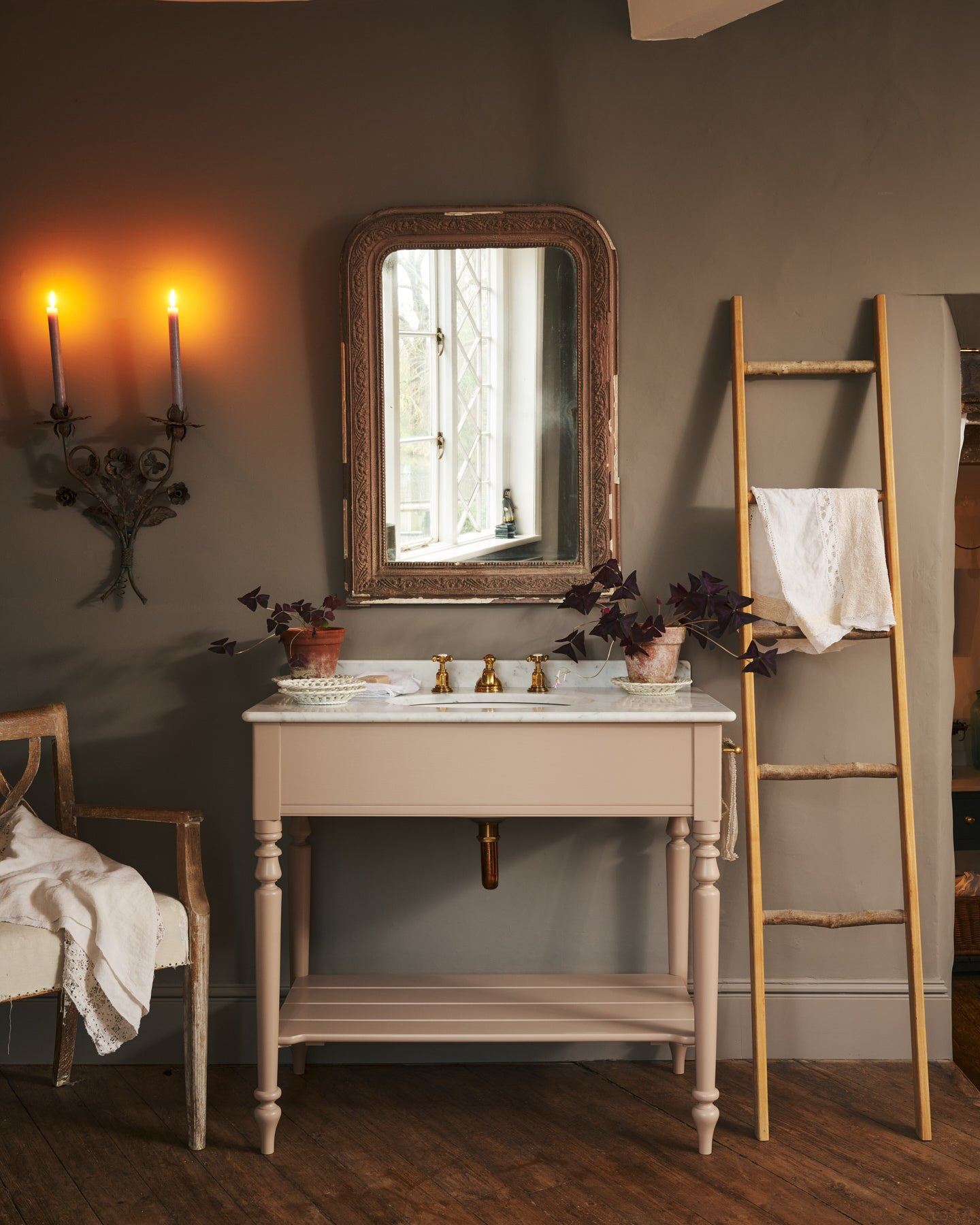 A straight-on view of a dusty-pink Victorian-inspired washstand with brass mixer taps in a greyish-brown painted room. The shot is atmospheric and moody, with two lit candles casting a subtle glow and light from an adjacent window illuminating the brass accents.