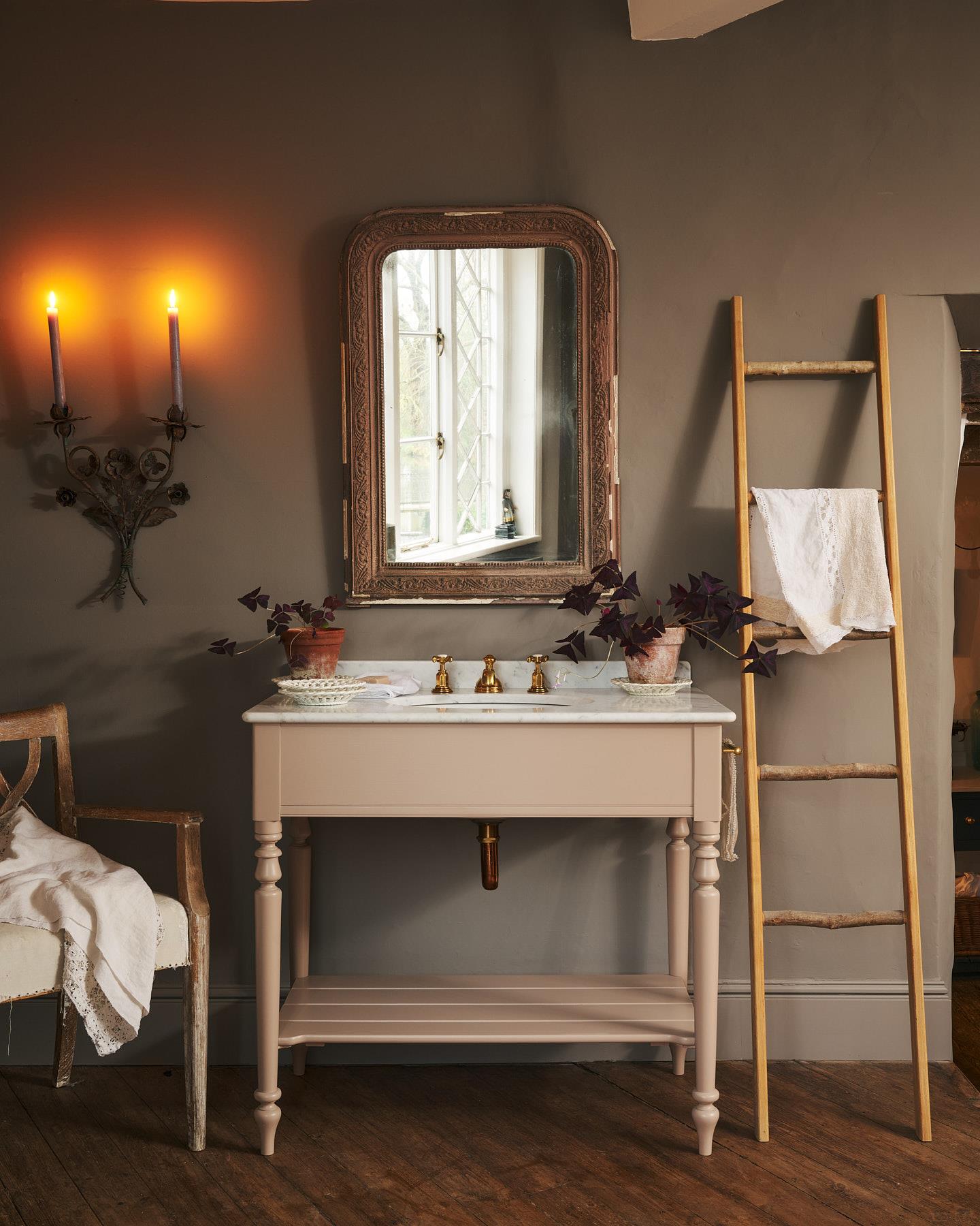 An oblique view of a dusty-pink Victorian-inspired washstand with brass mixer tap in a greyish-brown painted room. The shot is atmospheric and moody, with two lit candles casting a subtle glow and light from an adjacent window illuminating the brass accents.