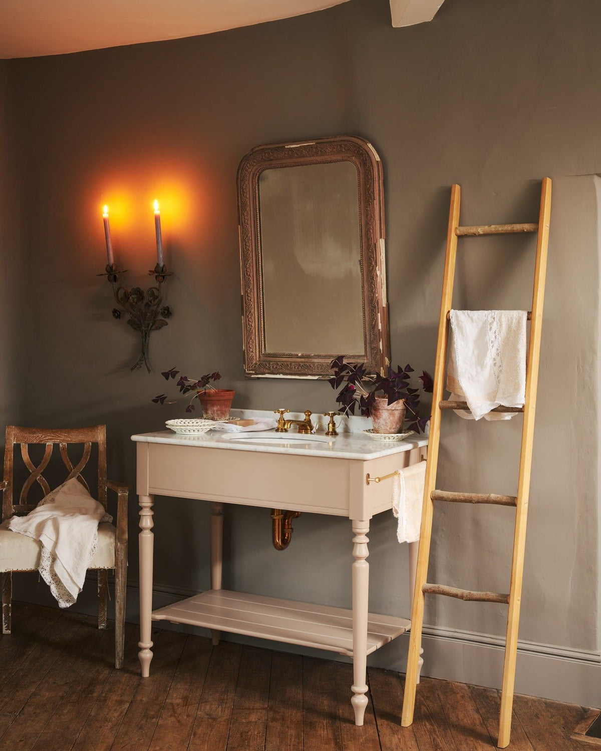 An oblique view towards a dusty-pink Victorian-inspired washstand with a brass mixer tap in a greyish-brown painted room. The shot is atmospheric and moody, with two lit candles casting a subtle glow and light from an adjacent window illuminating the brass accents. TEST
