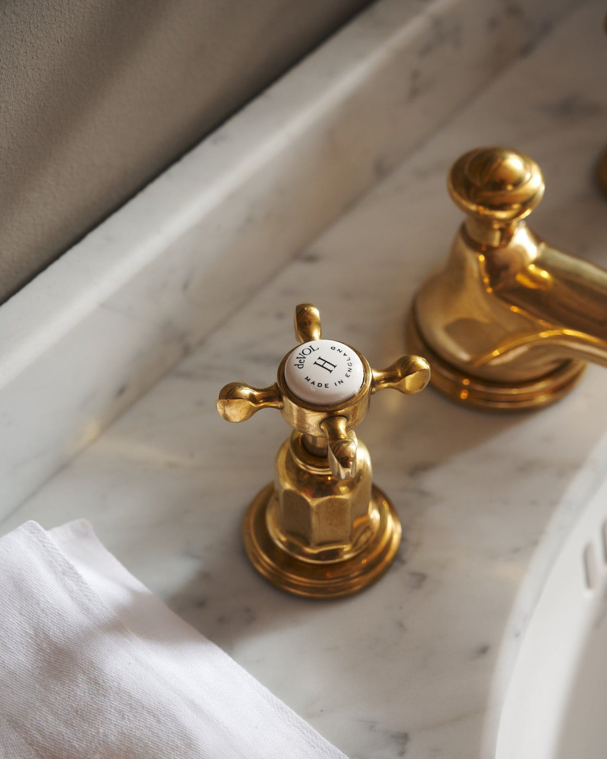 A close-up oblique view of the crosshead handle of an aged brass mixer tap for a bathroom basin sink. The letter 'H' is in the centre of the handle top, surrounded by the words 'deVOL, Made in England'. TEST