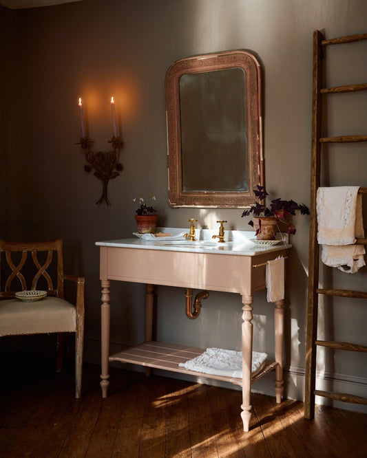 An oblique view towards a dusty-pink Victorian-inspired washstand with brass pillar taps in a greyish-brown painted room. The shot is atmospheric and moody, with two lit candles casting a subtle glow and light from an adjacent window illuminating the brass accents. | Large Princelet Pink shown