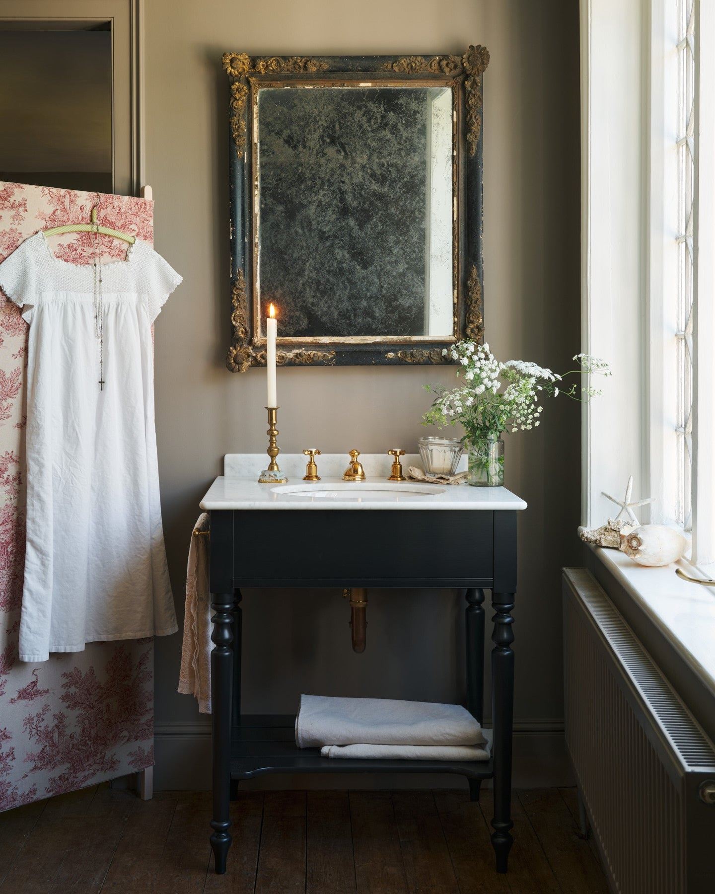 A straight-on view of a black Victorian-inspired washstand with a brass mixer tap in a greyish-brown painted room. A mottled antique mirror sits above the sink, an antique cotton dress hangs from a decorated partition and light streams through a nearby leaded window.