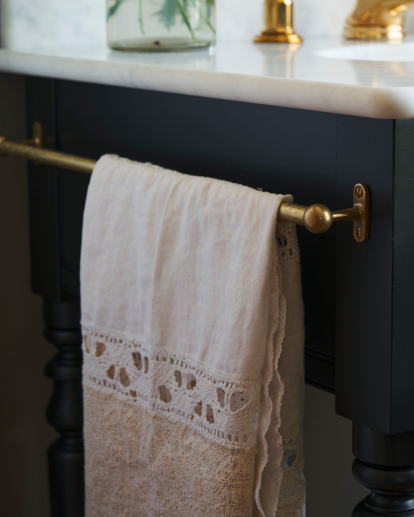 A detailed view of an aged brass towel rail attached to the left side of a Victorian-inspired washstand, a vintage lace hand towel hangs from the rail.