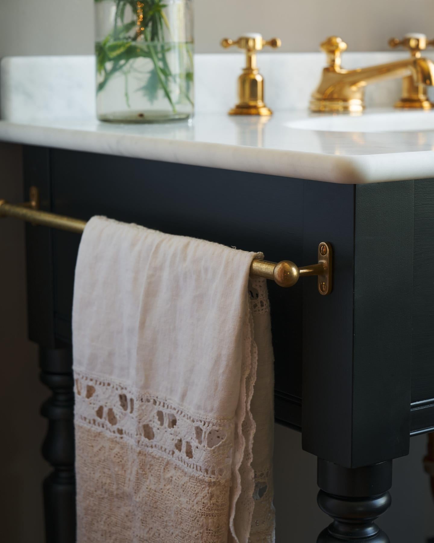 A detailed view of an aged brass towel rail attached to the left side of a Victorian-inspired washstand, a vintage lace hand towel hangs from the rail.