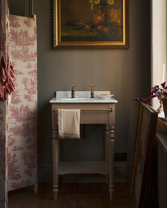 A straight-on view of a dusty-pink Victorian-inspired washstand with brass pillar taps in a greyish-brown painted room. The shot is atmospheric and moody, with an antique patterned partition and still-life oil paintings in gilded frames.