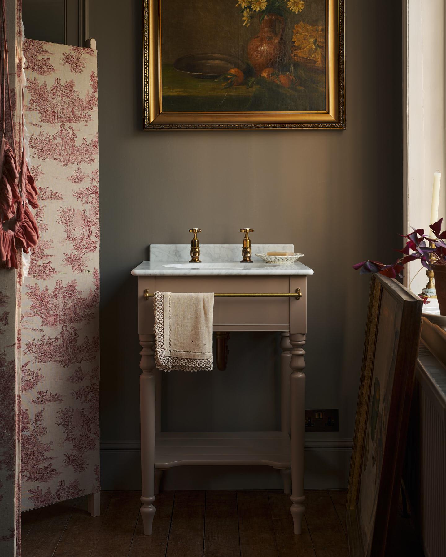 A straight-on view of a dusty-pink Victorian-inspired washstand with brass pillar taps in a greyish-brown painted room. The shot is atmospheric and moody, a vintage lace towel hangs from a brass rail attached to the front of the washstand.