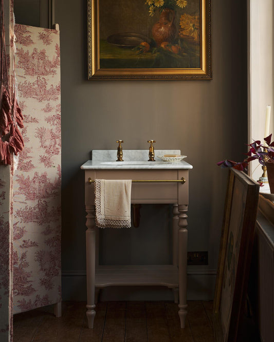A straight-on view of a mushroom-grey Victorian-inspired washstand with a brass mixer tap in a duck-egg blue painted room. The room is classic and bohemian with an ornate white mirror and elegant painted flowers across the top of the walls. | Princelet Pink with Pillar Taps shown
