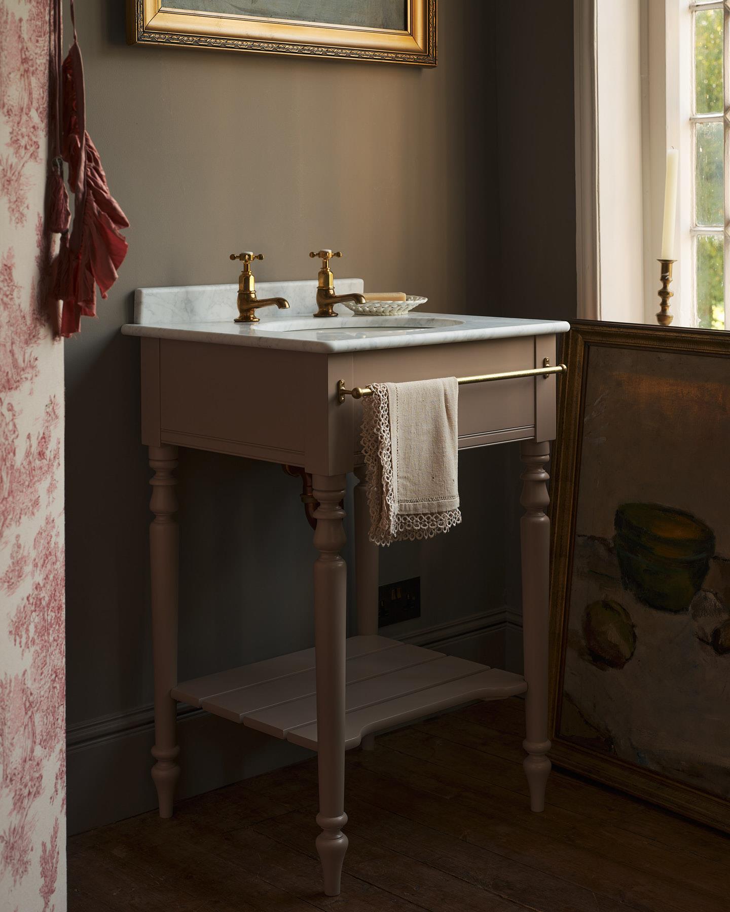 A straight-on view of a dusty-pink Victorian-inspired washstand with a brass mixer tap in a greyish-brown painted room. The shot is atmospheric and moody, with two lit candles casting a subtle glow and light from an adjacent window illuminating the brass accents.