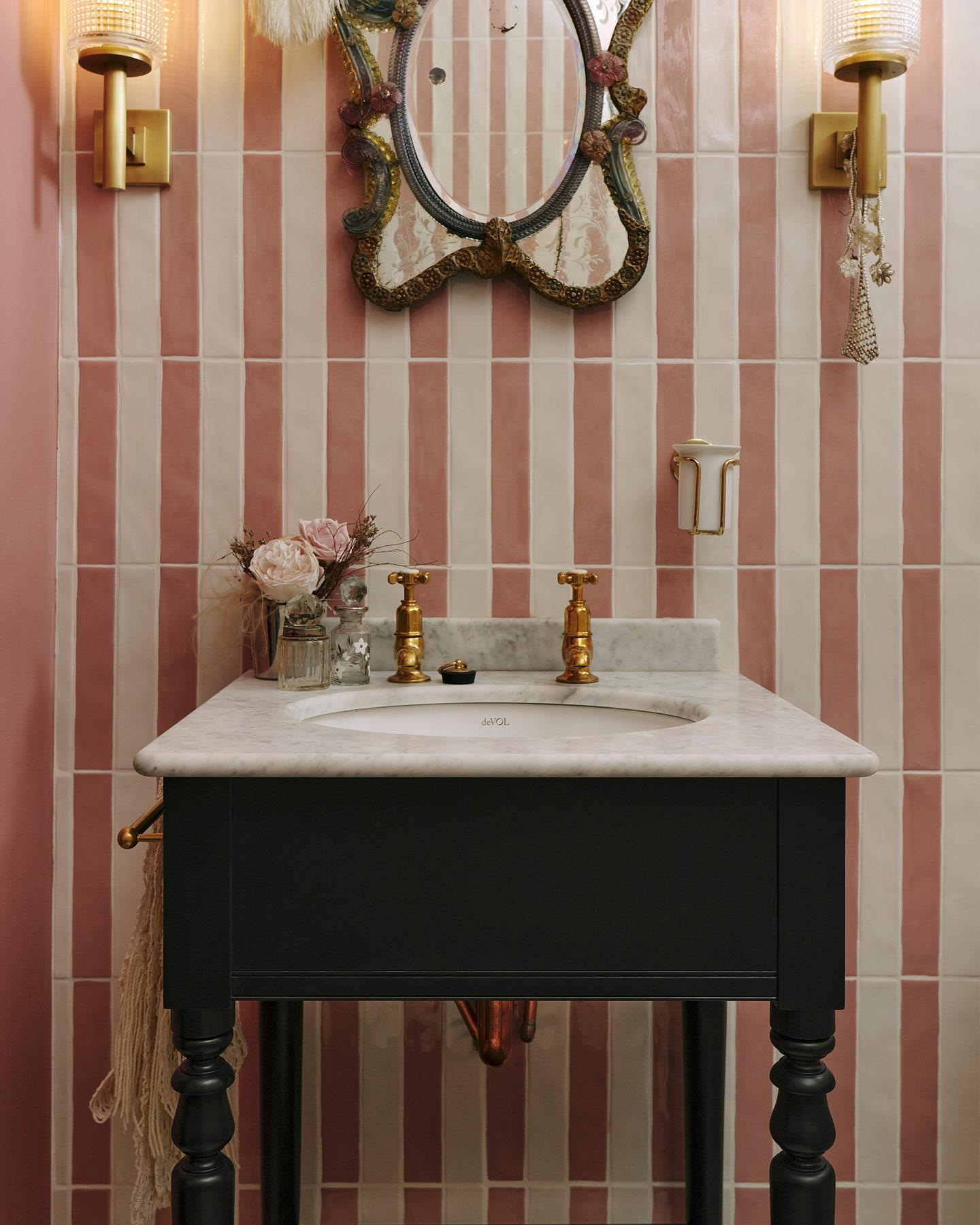 A straight-on view of a black Victorian-inspired washstand with a brass pillar tap against a pink and white splashback tiles. A decorative antique mirror sits above the sink, along with deco-style scones, and a brass cup holder on the wall.