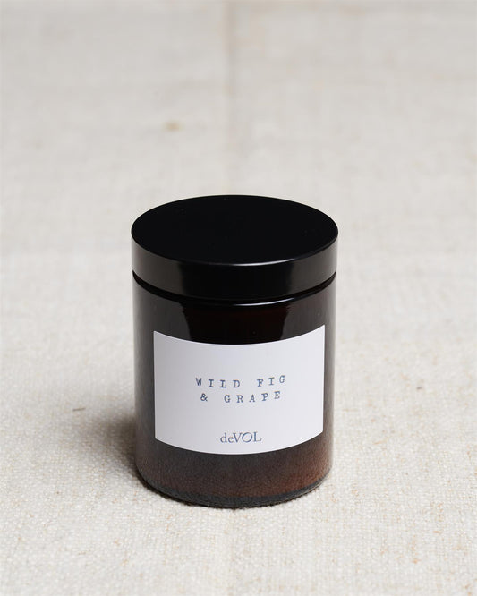 A medium scented candle in a brown glass jar with a black lid, the label reads 'Amber & Tonka Bean, deVOL'.