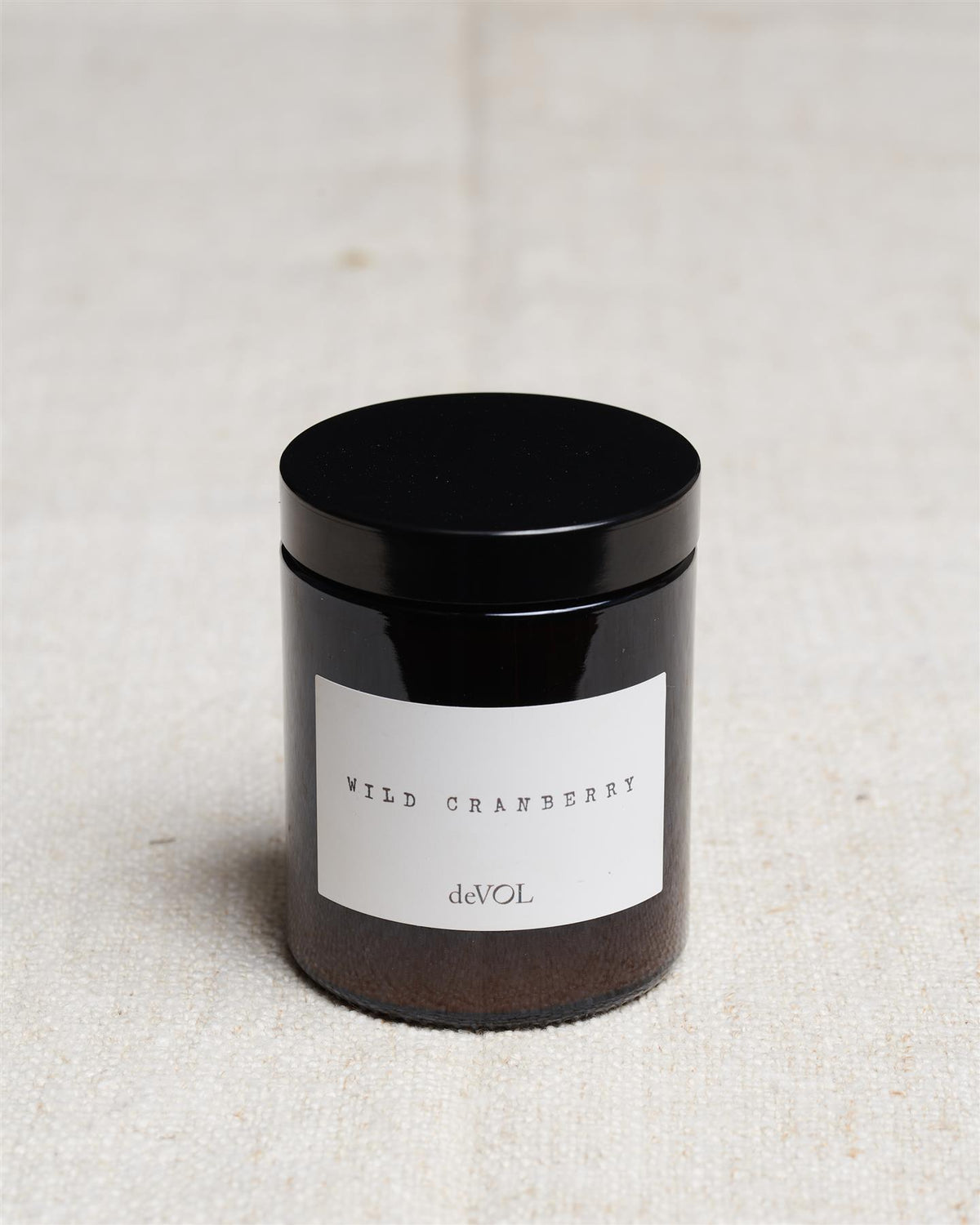 A medium scented candle in a brown glass jar with a black lid, the label reads 'Amber & Tonka Bean, deVOL'. TEST