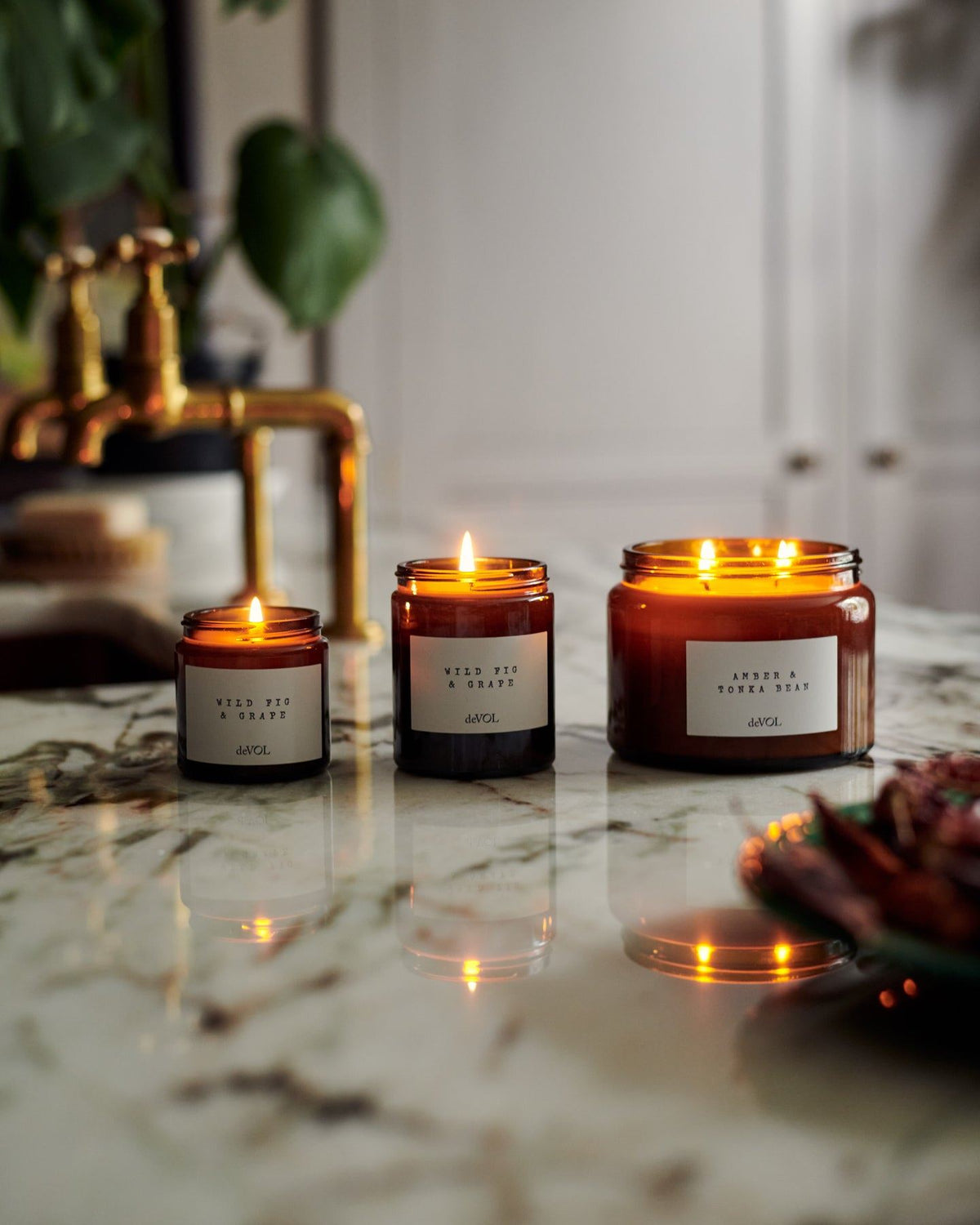 A lit small scented candle in a brown glass jar with one wick, in a vintage cupboard alongside other scented candles. TEST
