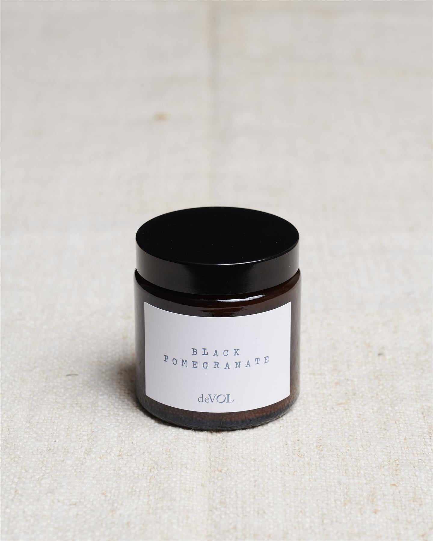 A small scented candle in a brown glass jar with a black lid, the label reads 'Amber & Tonka Bean, deVOL'.