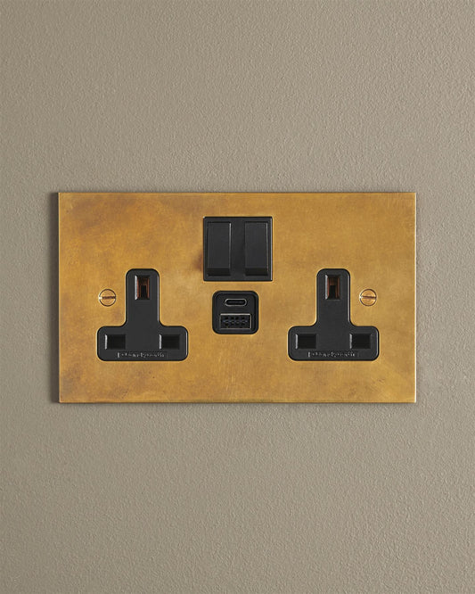 An Heirloom Brass Twin Plug Socket with USB A/C ports on a beige wall.