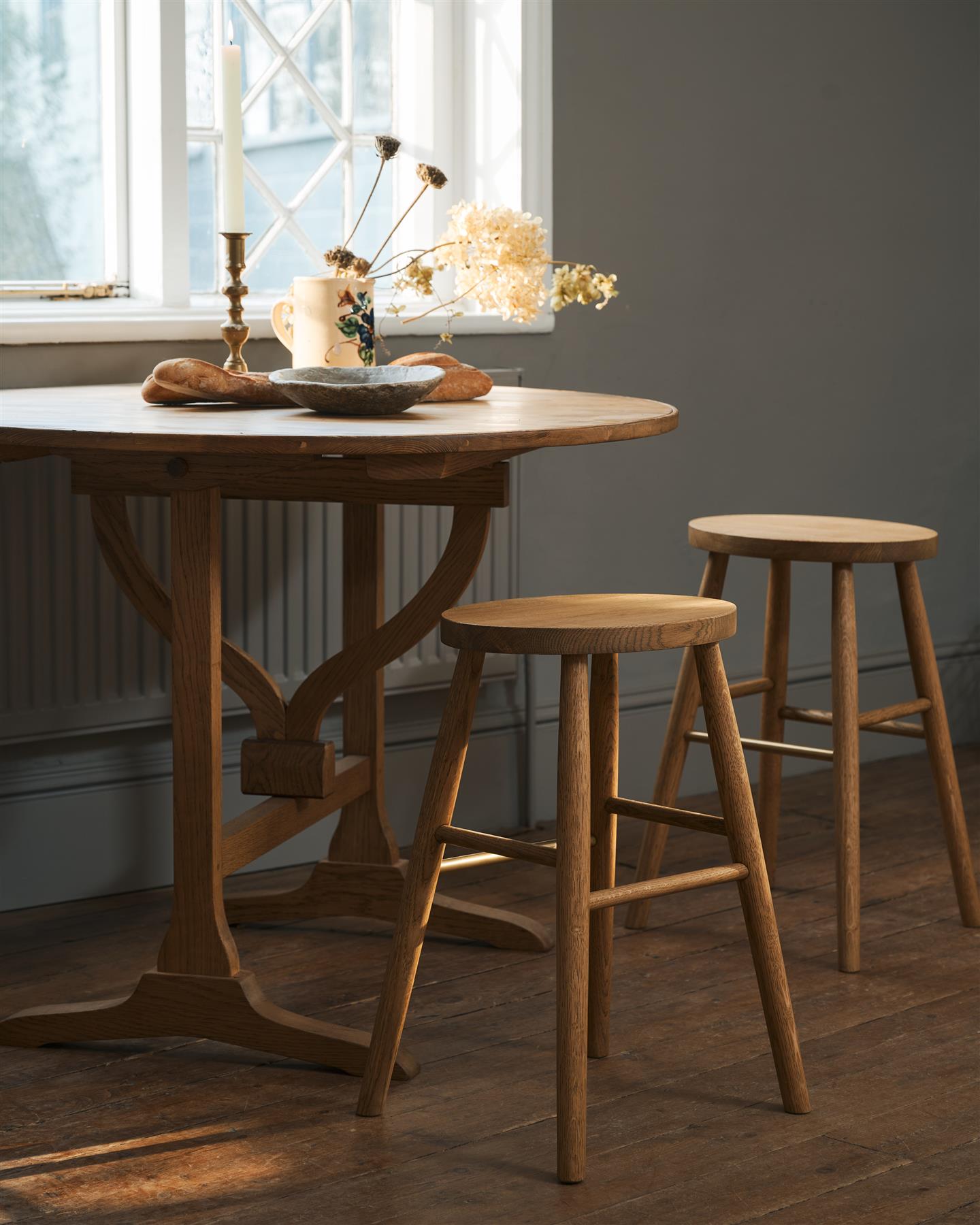 An oblique view of two simple oak milking stools with oval-shaped seats and brass footrails by a round pine table in a dark room.
