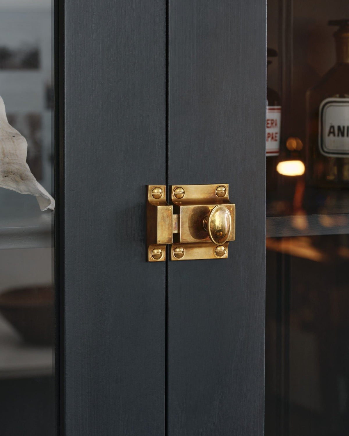 An oblique view of a classic-style square cupboard catch with an ornate oval-shaped knob in a warm burnished brass finish fitted to dark blue painted glazed cupboard doors. TEST