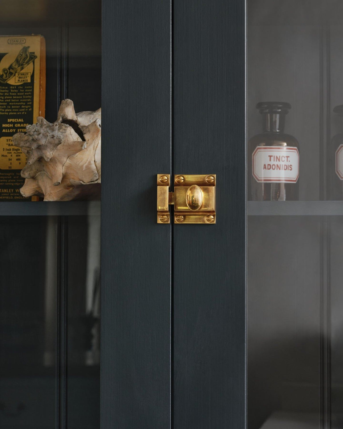 A straight-on view of a classic-style square cupboard catch with an ornate oval-shaped knob in a warm burnished brass finish fitted to dark blue painted glazed cupboard doors. TEST