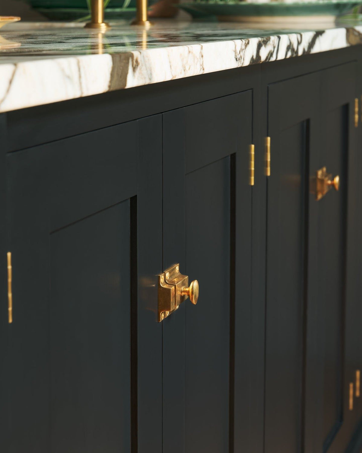 An oblique view of classic-style square cupboard catches in a warm burnished brass finish fitted to black painted kitchen cupboard doors. TEST