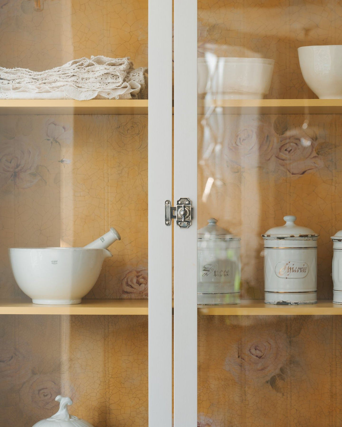 A straight-on view of a Victorian-inspired rack and pinion cupboard catch in a tarnished silver finish, fitted to a tall glazed cupboard with white painted doors and a yellow painted interior. TEST