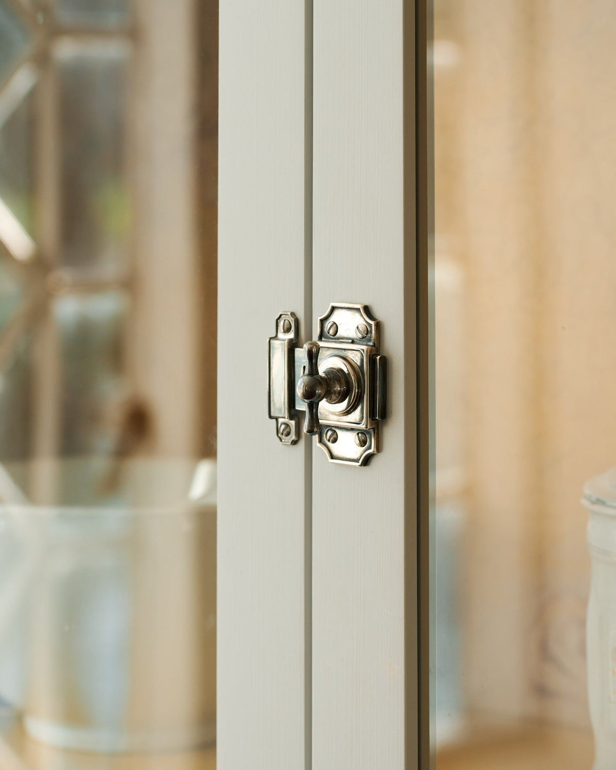 A close-up oblique view of a Victorian-inspired rack and pinion cupboard catch in a tarnished silver finish fitted to white painted doors. TEST