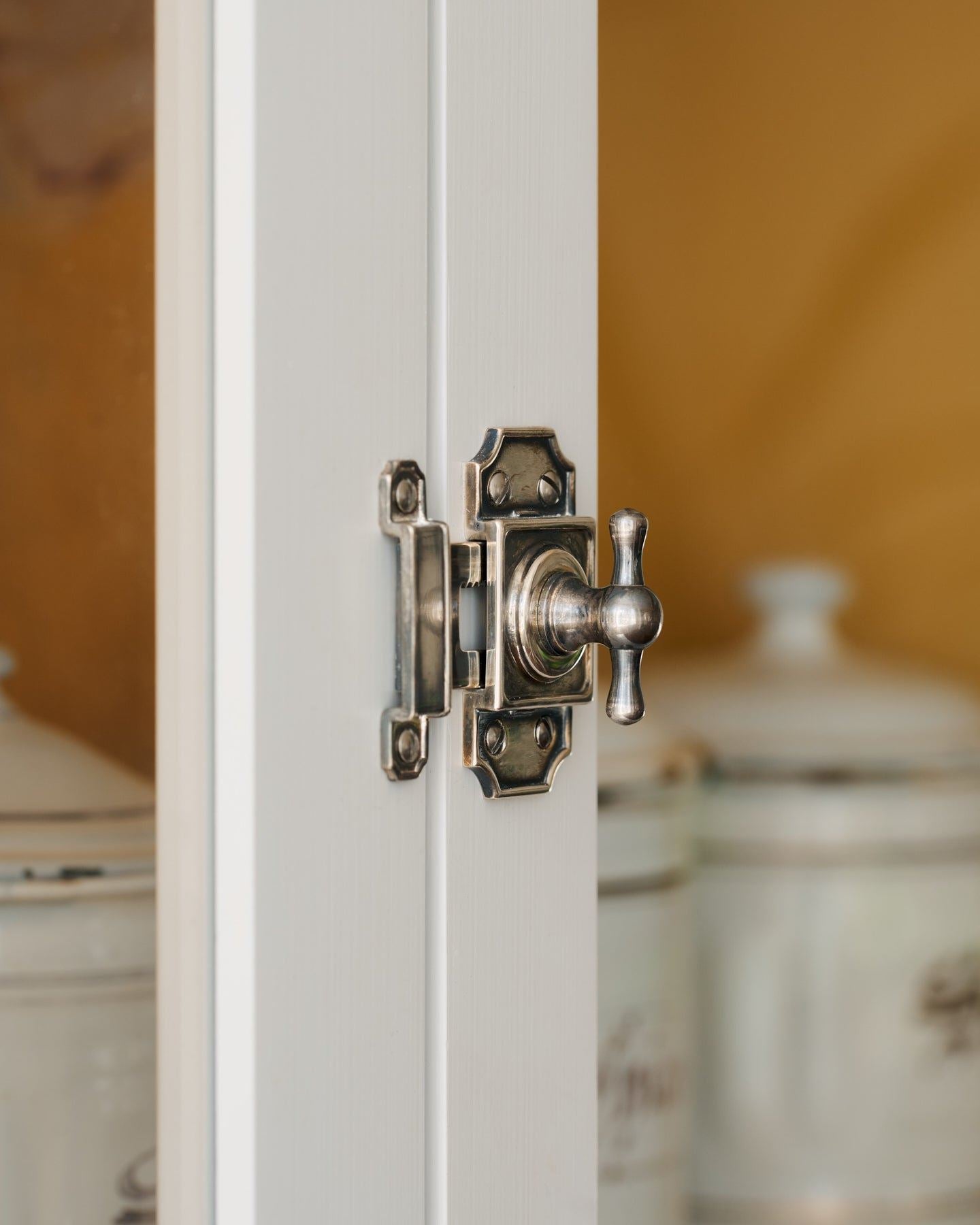 A close-up oblique view of a Victorian-inspired rack and pinion cupboard catch in a tarnished silver finish fitted to white painted doors.