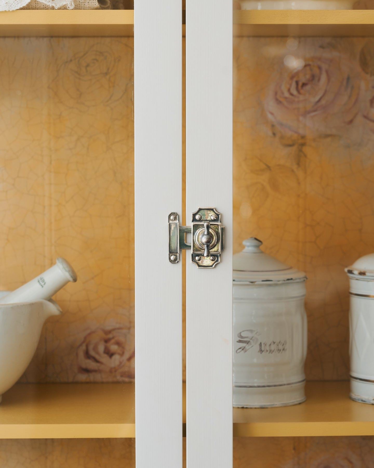 A close-up straight-on view of a Victorian-inspired rack and pinion cupboard catch in a tarnished silver finish, fitted to a tall glazed cupboard with white painted doors and a yellow painted interior.