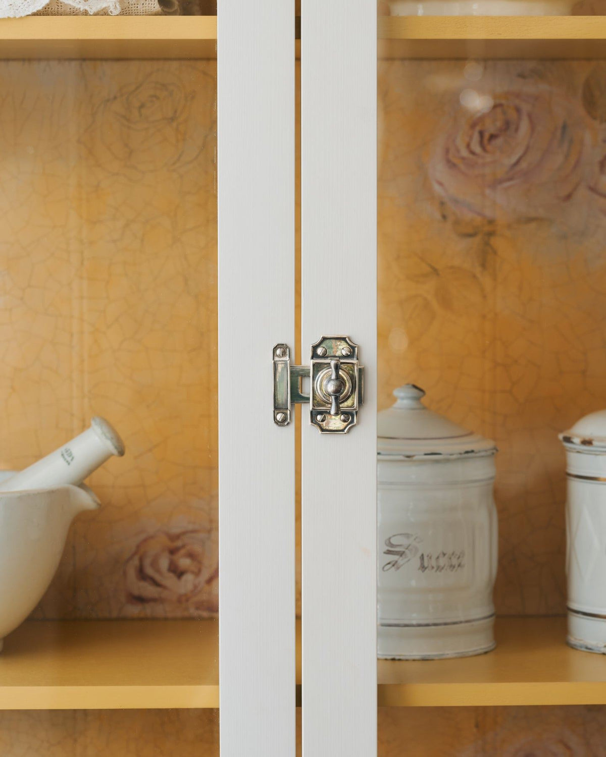 A close-up straight-on view of a Victorian-inspired rack and pinion cupboard catch in a tarnished silver finish, fitted to a tall glazed cupboard with white painted doors and a yellow painted interior. TEST