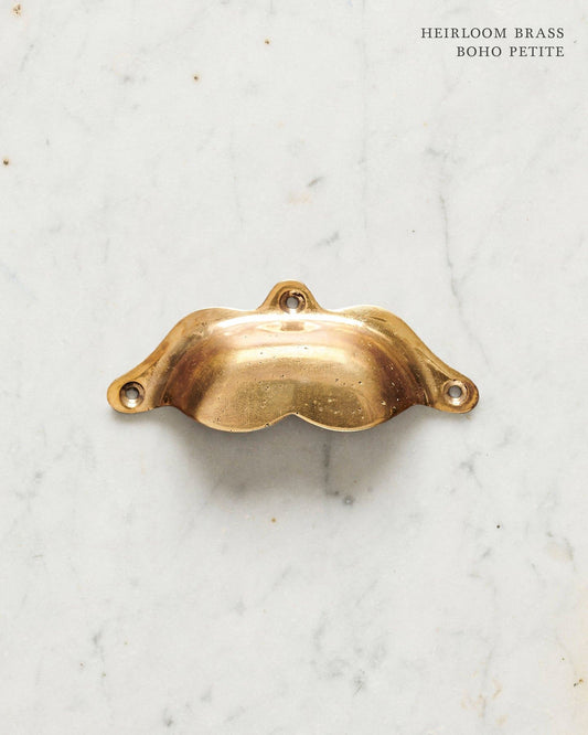 A small bohemian-style cup handle for a kitchen cupboard or drawer with a warm burnished brass finish.