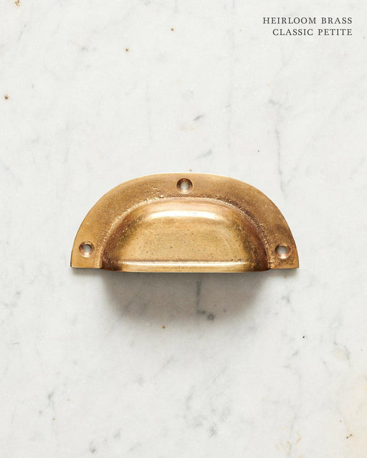 A small classic cup handle for a kitchen cupboard or drawer with a warm burnished brass finish.