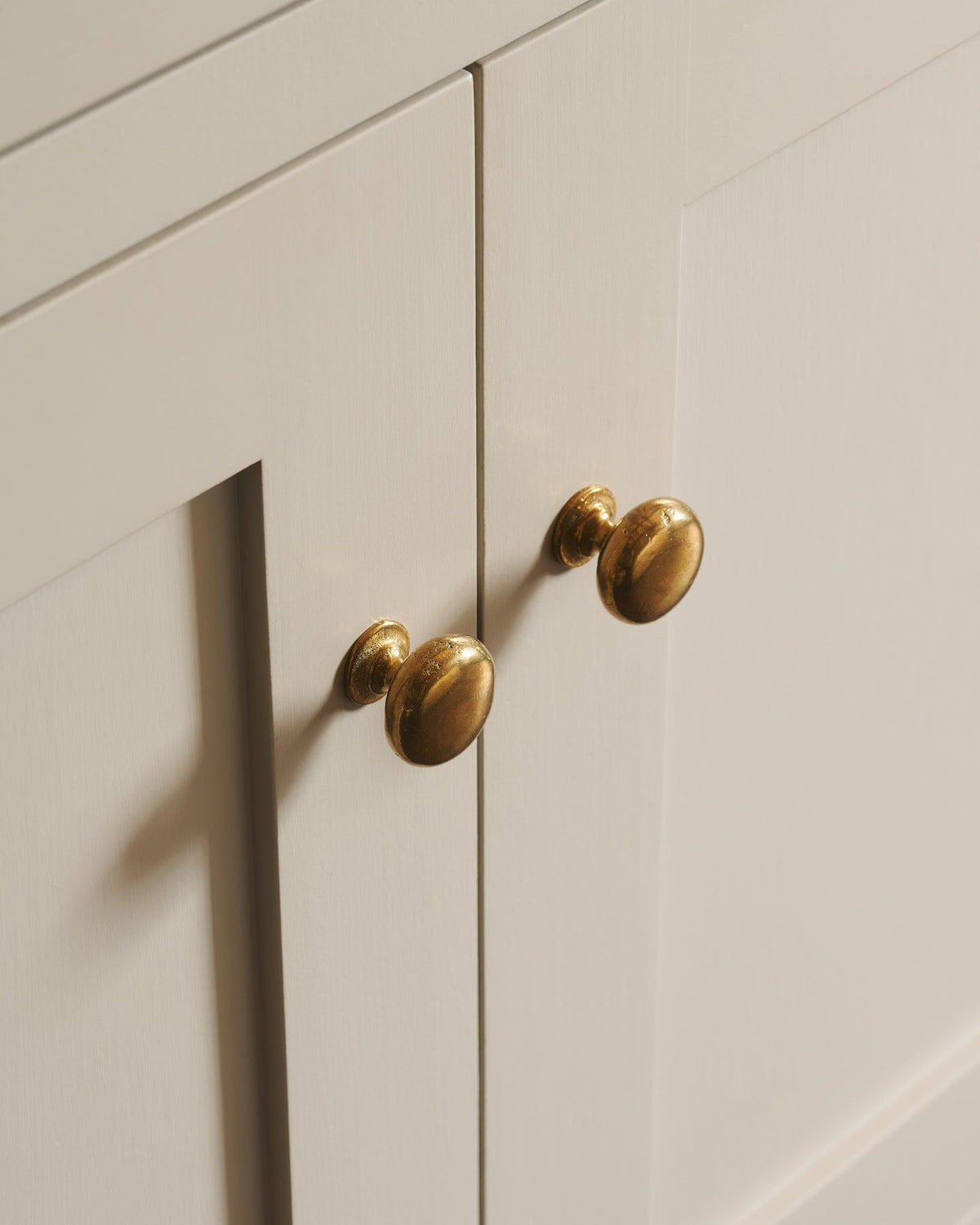 An oblique view of a pair of classic knobs in a warm burnished brass finish, fitted to a mushroom painted Shaker-style kitchen cupboard.|Heirloom Brass Medium shown TEST