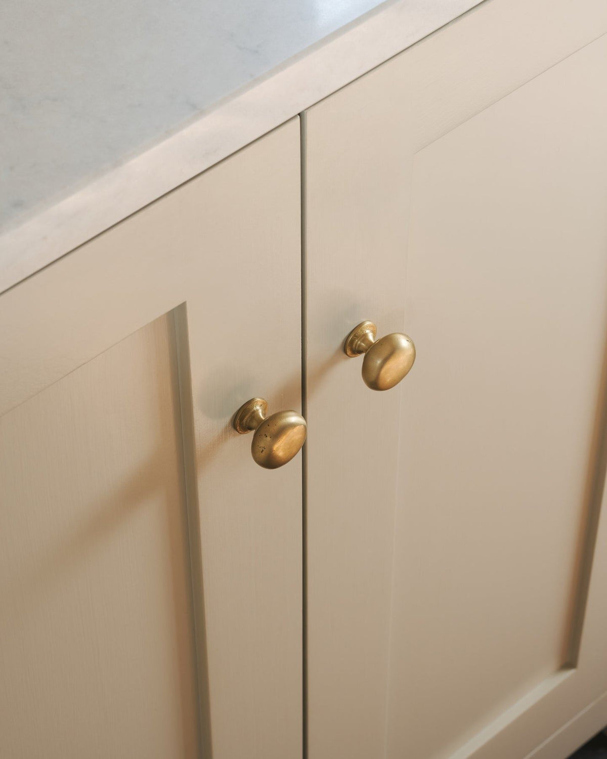 An oblique view of a pair of classic knobs in an aged brass finish, fitted to a mushroom painted Shaker-style kitchen cupboard. TEST