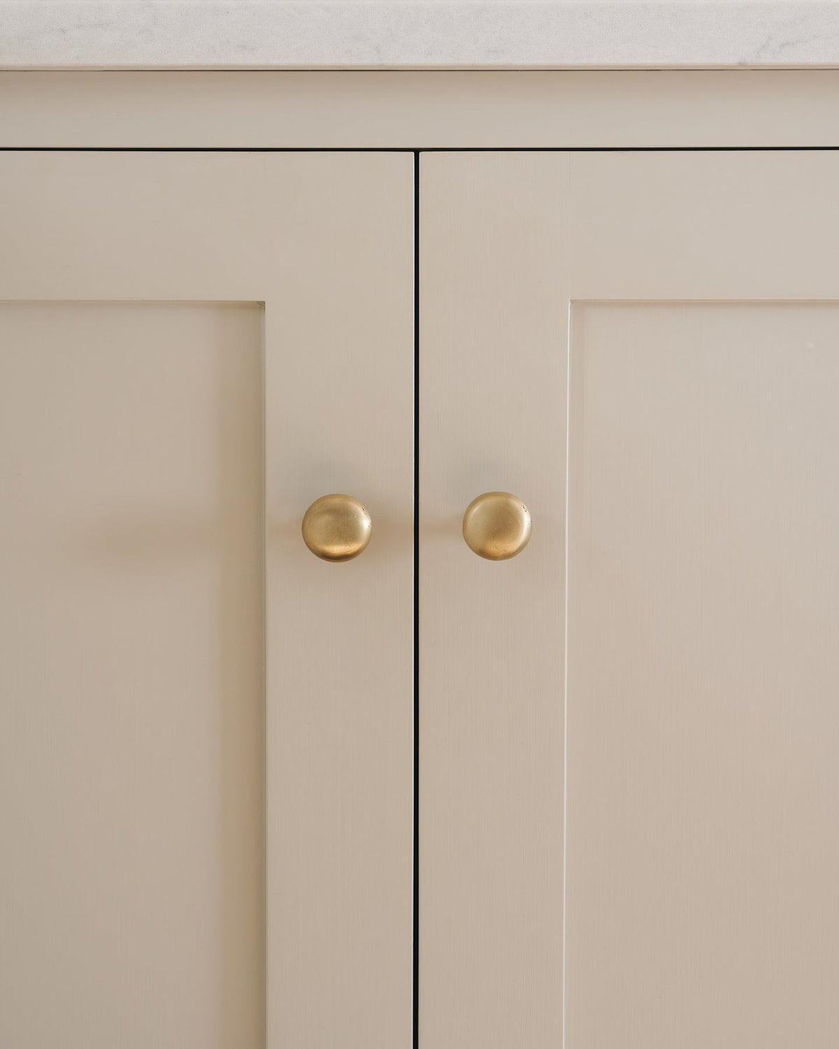 A straight-on view of a pair of classic knobs in an aged brass finish, fitted to a mushroom painted Shaker-style kitchen cupboard. TEST