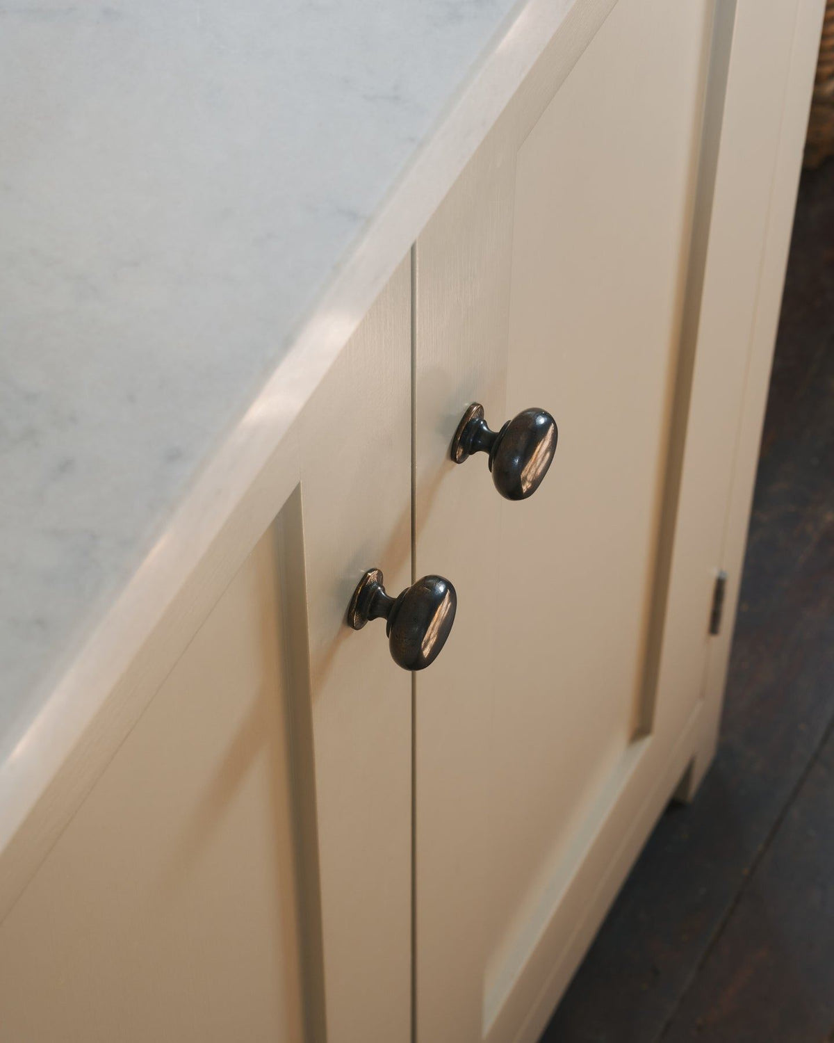 A close-up oblique view of a pair of classic knobs in a dark oxidised bronze finish, fitted to a mushroom painted Shaker-style kitchen cupboard. TEST