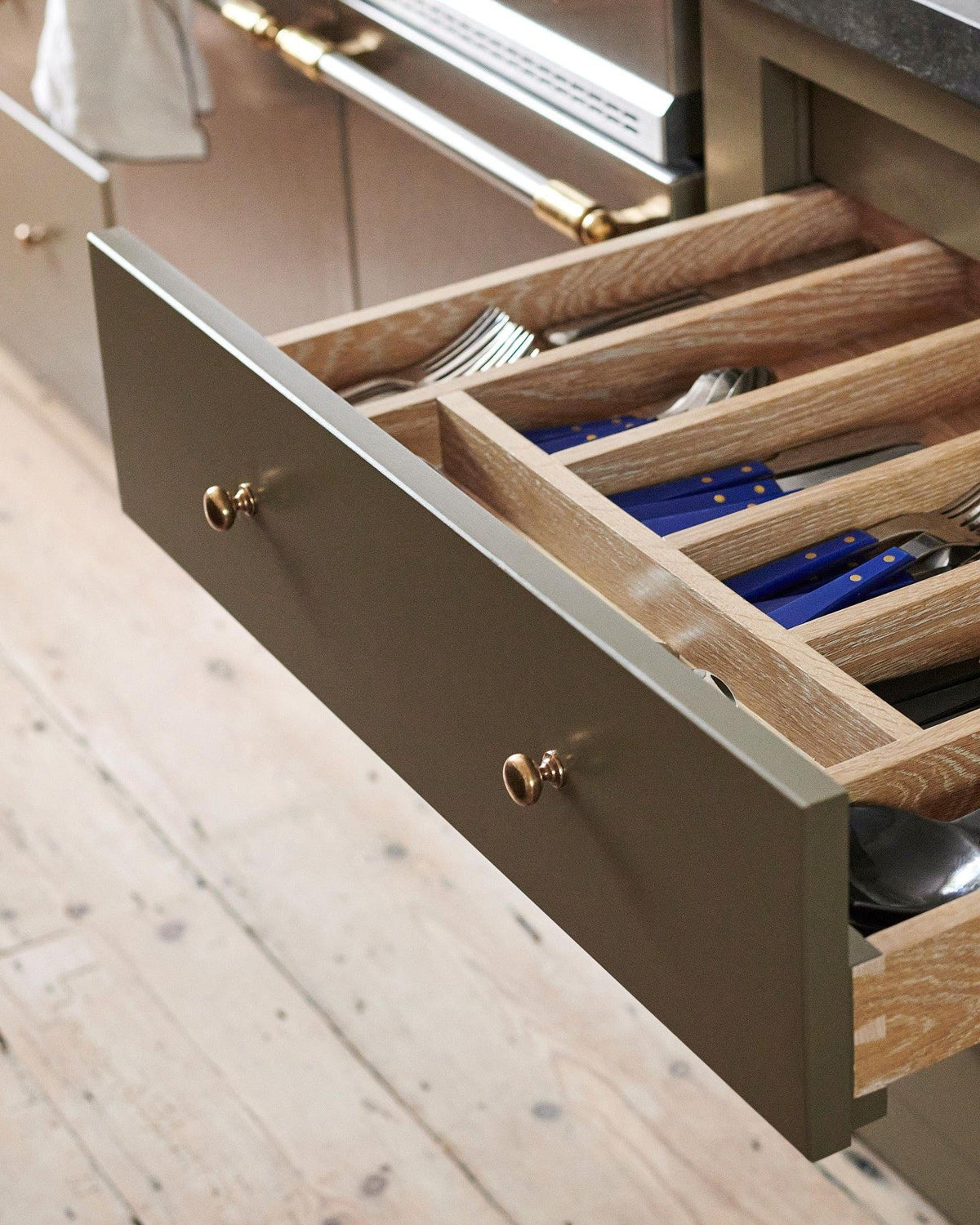 An oblique view of a mossy green painted open cupboard drawer with classic knobs in a bright polished bronze finish and an oak cutlery tray full of utensils.|Polished Bronze Medium shown TEST
