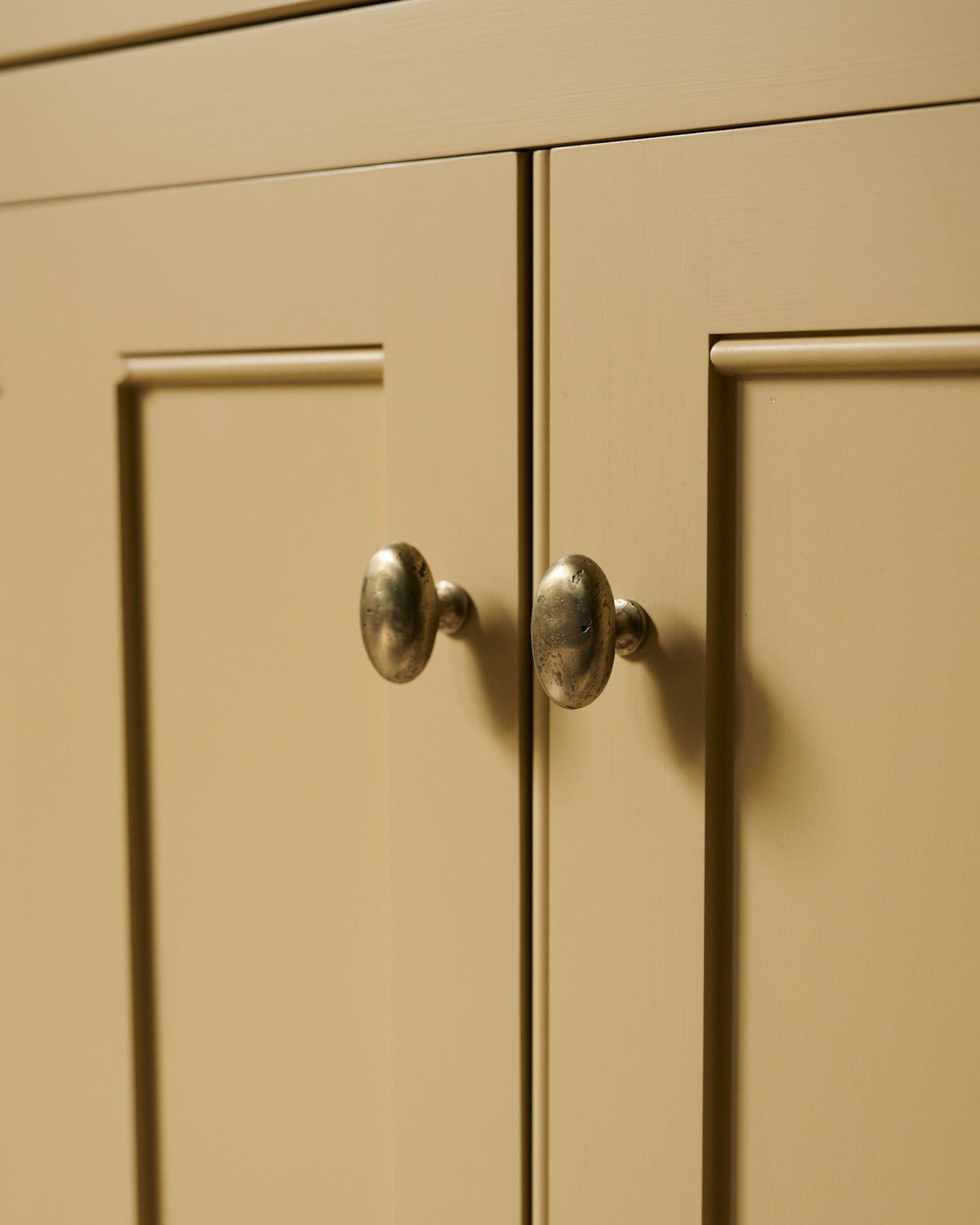 An oblique view of a pair of oval-shaped knobs in an aged brass finish, fitted to a yellow painted Georgian-style kitchen cupboard. | Large shown
 TEST